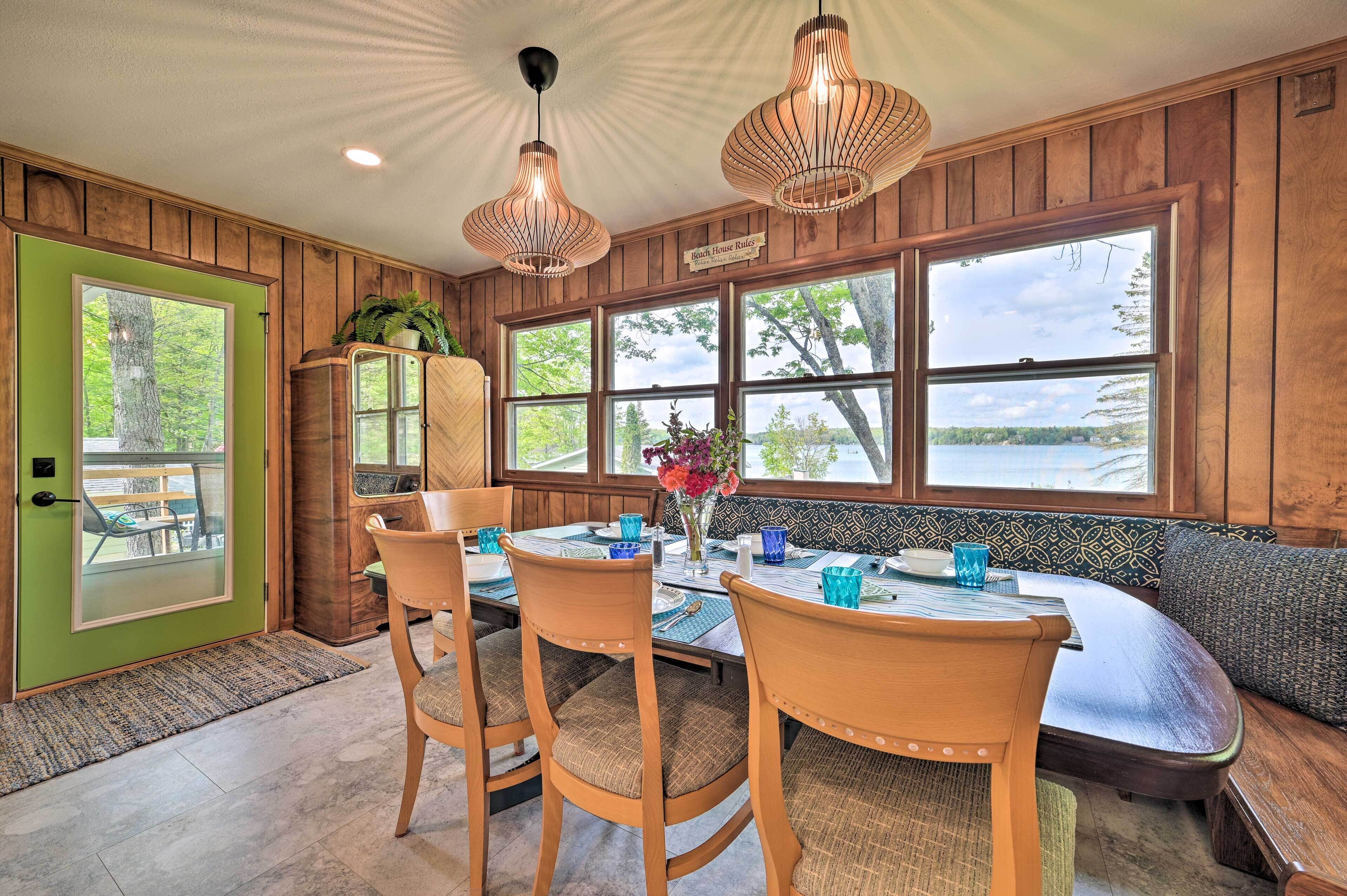 A dining area with a wooden table, chairs, and a bench. There are large windows with a view of trees and a lake.