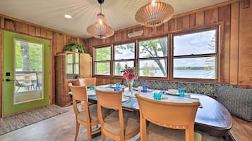 A dining area with a wooden table, chairs, and a bench. There are large windows with a view of trees and a lake.