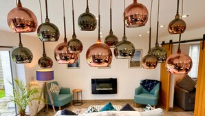 A modern living room with multiple hanging copper pendant lights, a large flat-screen TV, and a comfortable seating area with a marble throw blanket. - Stylish, Dog Friendly Coastal Bungalow With Garden (Holyhead)