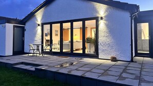 A modern house with a large glass door leading to a well-lit interior, a tiled patio, and a small outdoor seating area. - Stylish, Dog Friendly Coastal Bungalow With Garden (Holyhead)