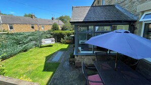 A patio area with a table and chairs, a swing, and a view of the garden and neighboring houses. - Acacia House  - Holiday Cottage Wolsingham (Wolsingham)
