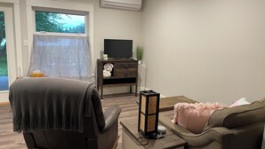 A living room with a sofa, a TV on a wooden stand, a small table with a lamp, and a window with a view of trees. - New 1 bedroom Sooke oceanfront guest suite  (Sooke)