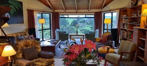 A cozy living room with a sofa, armchairs, a coffee table, and a bookshelf. Large windows offer a view of greenery outside. - Balcony river views and great value (Haruru)