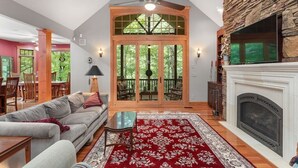 A spacious living room with a fireplace, a large window with a view of greenery, a ceiling fan, and a red patterned rug. - Large Luxury Home w/Hot Tub & Game Room with access to Rumbling Bald Resort (Lake Lure)