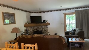 A cozy living room with a stone fireplace, a television mounted above it, a comfortable sofa, a wooden dining table, and a small side table with decorative items. - Newer Rustic WiFi Villa by Starved Rock sleeps 7. A guest favorite!! (Utica)