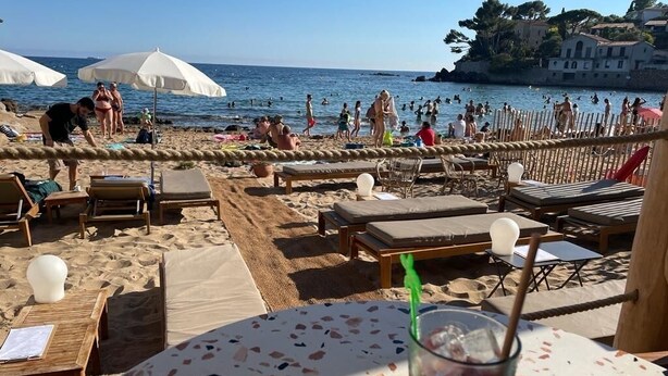 Una playa con tumbonas y sombrillas, una mesa con una bebida, y vista al mar y edificios al fondo.