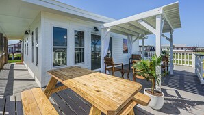 A wooden deck with a picnic table and chairs, a potted plant, and a white building with windows and a covered area. - The Surfsonian - 2nd Row Beach House - Dog Friendly - Ocean View (Freeport)