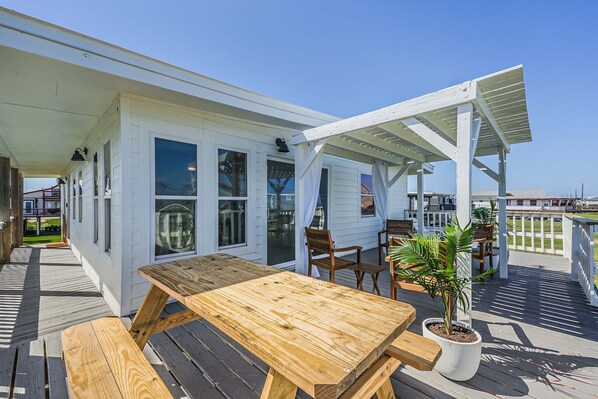 A wooden deck with a picnic table and chairs, a potted plant, and a white building with windows and a covered area.