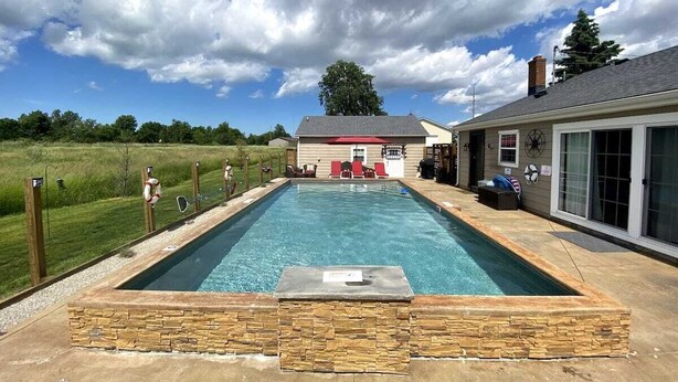 Une piscine rectangulaire bordée de pierres, entourée d’une pelouse et d’une maison avec un toit rouge et un revêtement extérieur blanc.