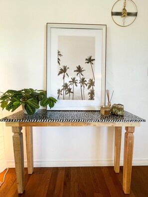 A small table with a patterned tabletop sits against a wall, featuring a framed picture, a potted plant, and a decorative candle holder.