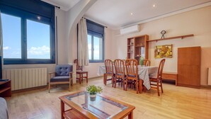 Een ruime woonkamer met een eethoek, grote ramen, een houten vloer en een moderne keuken op de achtergrond.