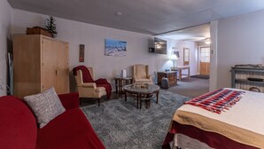 En hyggelig stue med en rød sofa, en beige lænestol, et glasbord og en stor seng med et rødt og hvidt tæppe. Der er et træskab, et lille bord med en lampe og et fjernsyn ophængt på væggen.