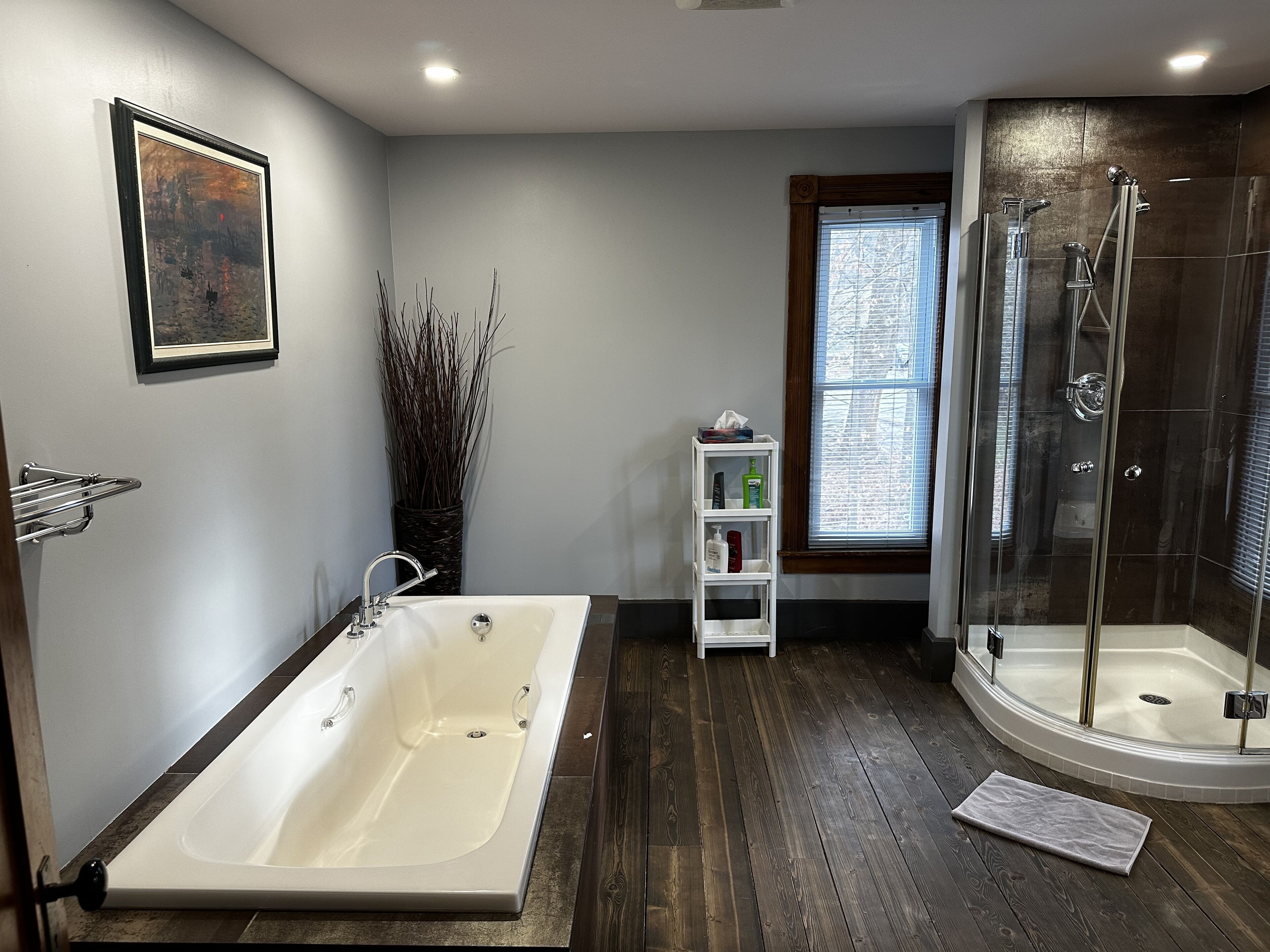 A bathroom with a bathtub, a glass-enclosed shower, a wooden floor, a small white shelf with toiletries, and a framed picture on the wall.