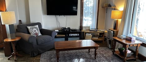 Una sala de estar con sofá, mesa de centro, televisor de pantalla plana, lámpara y ventana con cortinas.
