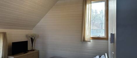 A bedroom with a sloped ceiling, a bed, a dresser, a television, and a window with curtains.