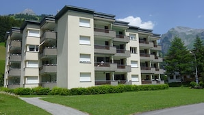 Un condominio a più piani con balconi, un vialetto lastricato e montagne sullo sfondo.