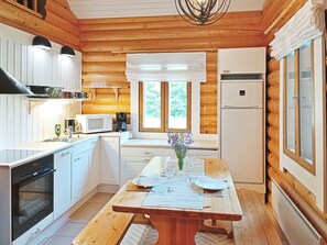 Cocina de cabaña de troncos con una mesa de comedor, armarios blancos, un refrigerador y una ventana con vista a la vegetación.