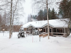 A log cabin with a snow-covered roof, surrounded by trees and a small Christmas tree.