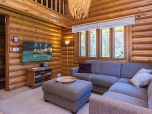 Een houten blokhut van binnen met een televisie, bank, voetenbankje en een raam met uitzicht op bomen.