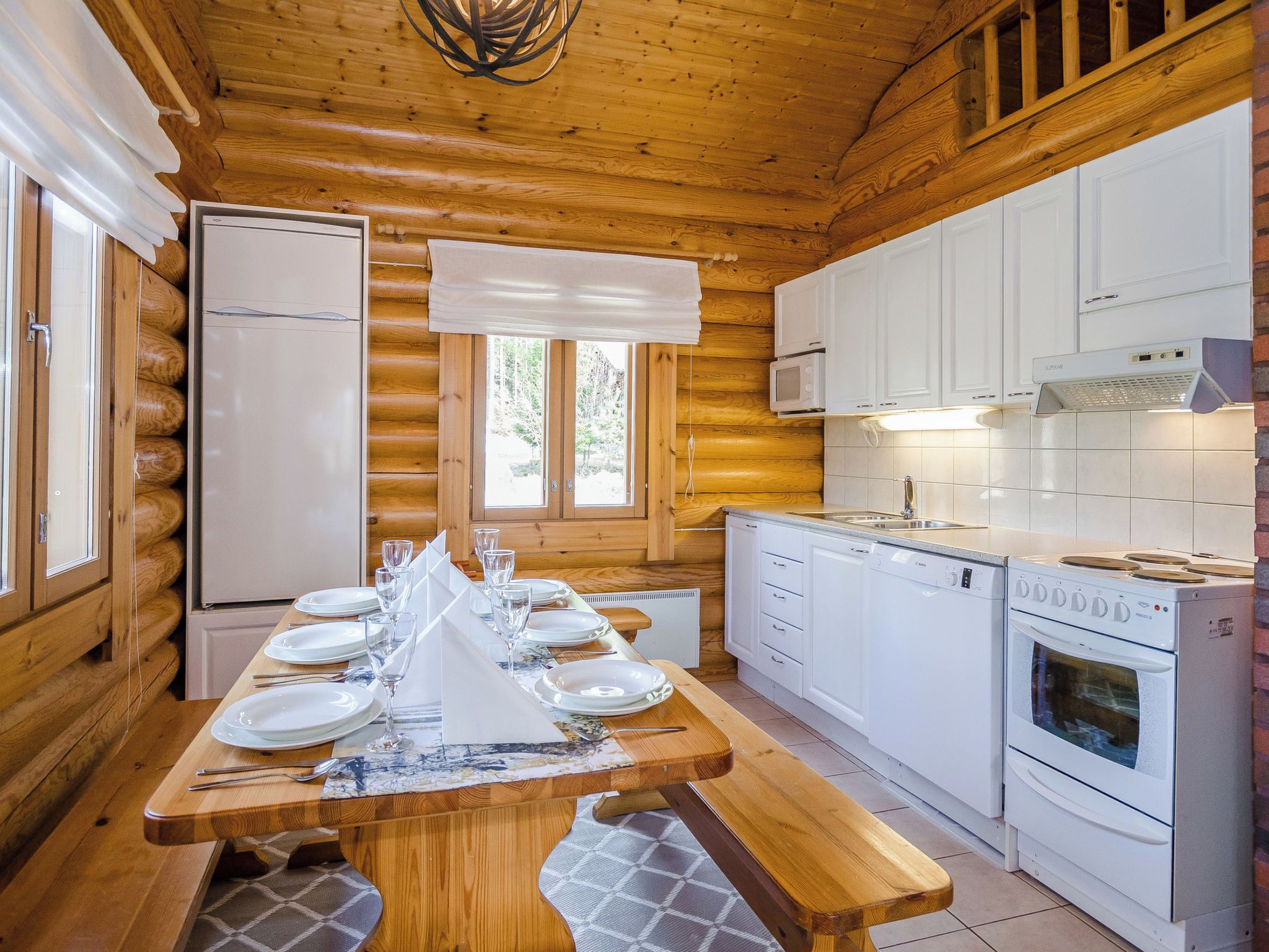 Interior de una cabaña de madera con una mesa de comedor para seis personas, una cocina con electrodomésticos blancos y una ventana con vistas a árboles.