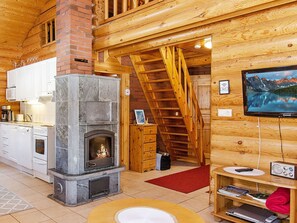 A cozy cabin interior with a fireplace, wooden walls, a kitchen area, and a staircase leading to an upper level.