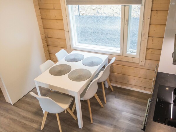 A small dining area with a white table and four chairs, a window with a view of a gravel path, and a kitchen area with a stove.
