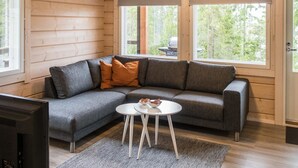 L’intérieur d’un chalet moderne en bois présente un canapé d’angle gris, une petite table blanche et une grande fenêtre donnant sur le paysage extérieur.