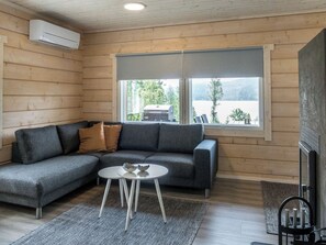 A modern log cabin interior with a grey sectional sofa, a small round table, and a large window offering a view of a lake and trees.