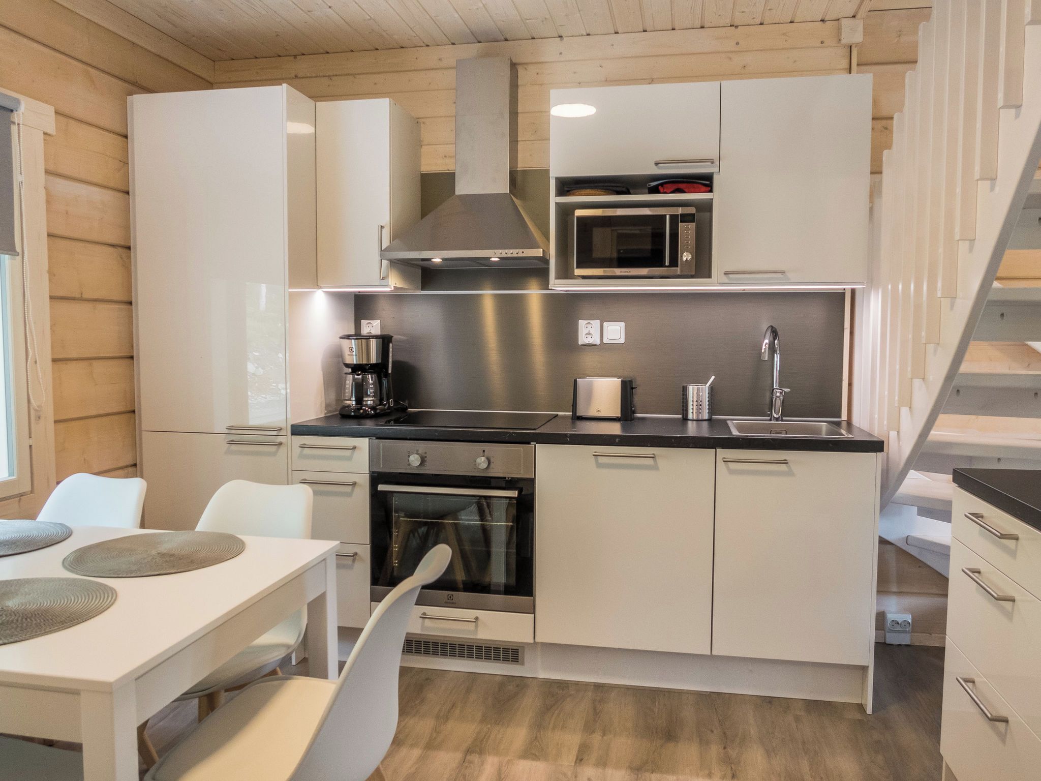 A compact kitchen with a microwave, stove, and coffee maker. There is a dining table with chairs and a staircase to the right.