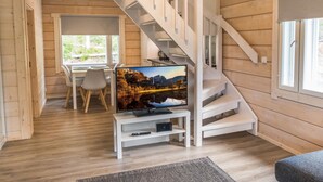 A modern log cabin interior with a wooden staircase, a dining area with a table and chairs, a television on a white stand, and a large window offering a view of the outdoors.