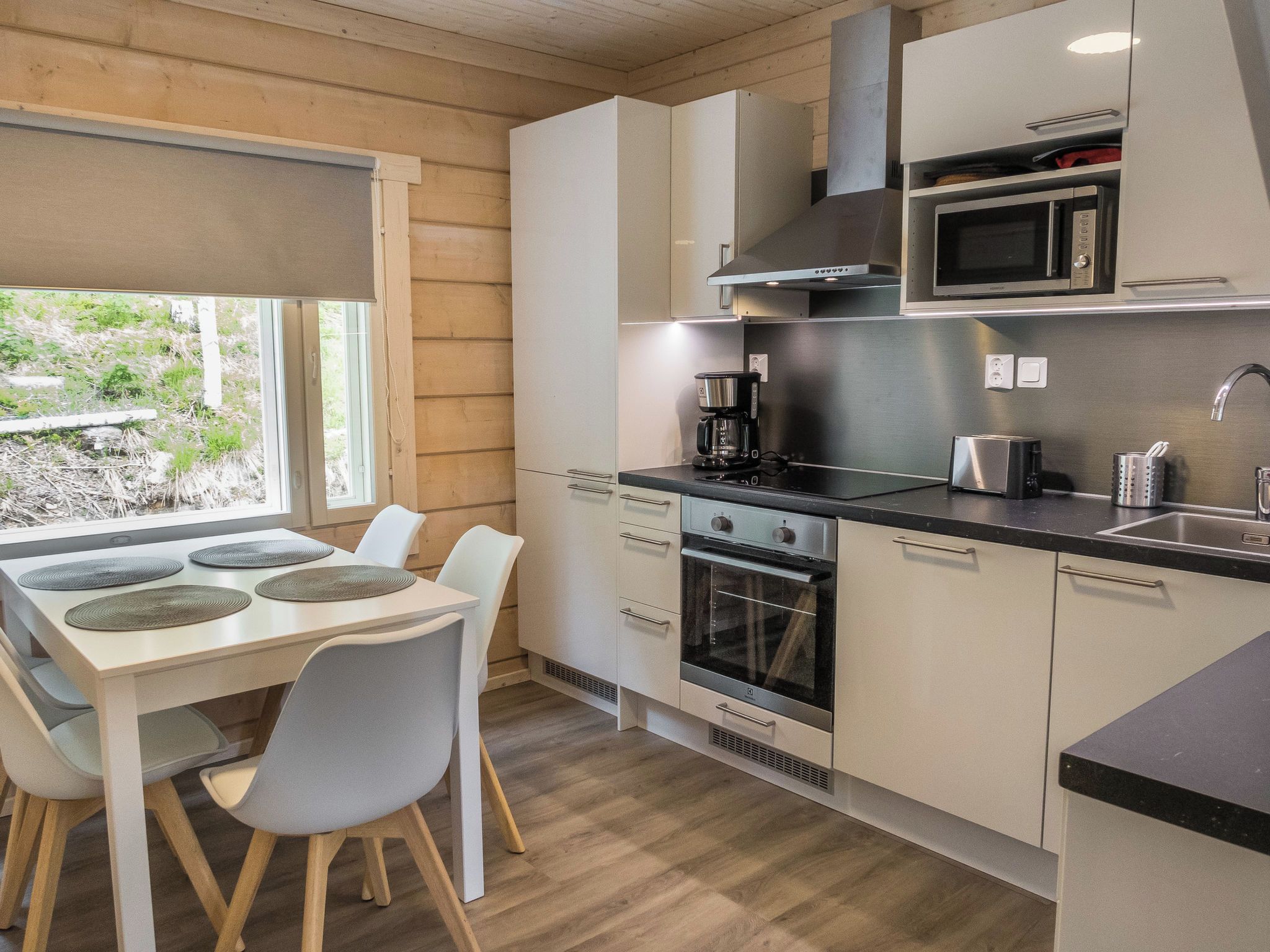 A modern kitchen with a dining area, featuring stainless steel appliances, white cabinetry, and a view of greenery outside.