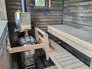 Puinen mökki, jonka sisätiloissa on puukamiina, penkki ja ikkuna.