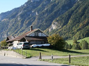 Ein traditionelles Chalet mit geneigtem Dach, Schornstein und kleinem Fenster. Vorne ist ein weißer Van geparkt.