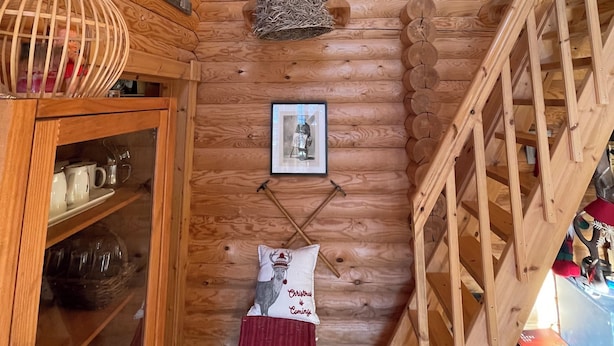 Interior de una cabaña de madera con una escalera, un cuadro enmarcado, un manto de piel blanca y una cesta.