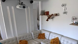 A modern living room with a light grey sectional sofa, two brown throw pillows, a ceiling-mounted projector, and a wall-mounted shelf with books and decorative items. - Comfortable family home in the mountains, near lake and resorts (La Rochette)