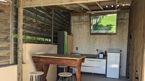 A rustic outdoor kitchen with a wooden table, two stools, a white refrigerator, and a window overlooking a green landscape. - Comfortable family home in the mountains, near lake and resorts (La Rochette)