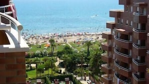 Un hotel frente a la playa con varios balcones, una playa al fondo y abundante vegetación.