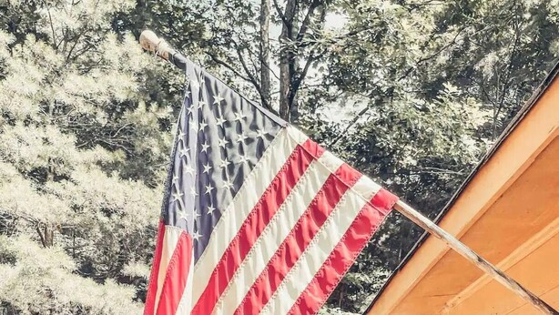 Un drapeau américain est accroché à l’extérieur d’un bâtiment dont la façade est en bois, et un jardin avec des arbustes verts se trouve à proximité.