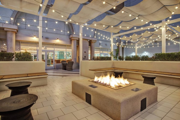 Een terras met buitenmeubilair, een vuurplaats, lichtjes en uitzicht op het interieur van het gebouw.