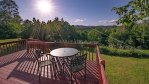 A wooden deck with a round table and chairs, surrounded by a wooden railing, overlooking a lush green landscape. - Amazing View, Walk to lake, very Central, borders state park. NEWLY RENOVATED! (Waterbury Center)