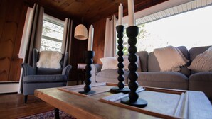 A cozy living room with a wooden ceiling, a grey sofa, a blue armchair, and a wooden coffee table with candles. - Amazing View, Walk to lake, very Central, borders state park. NEWLY RENOVATED! (Waterbury Center)