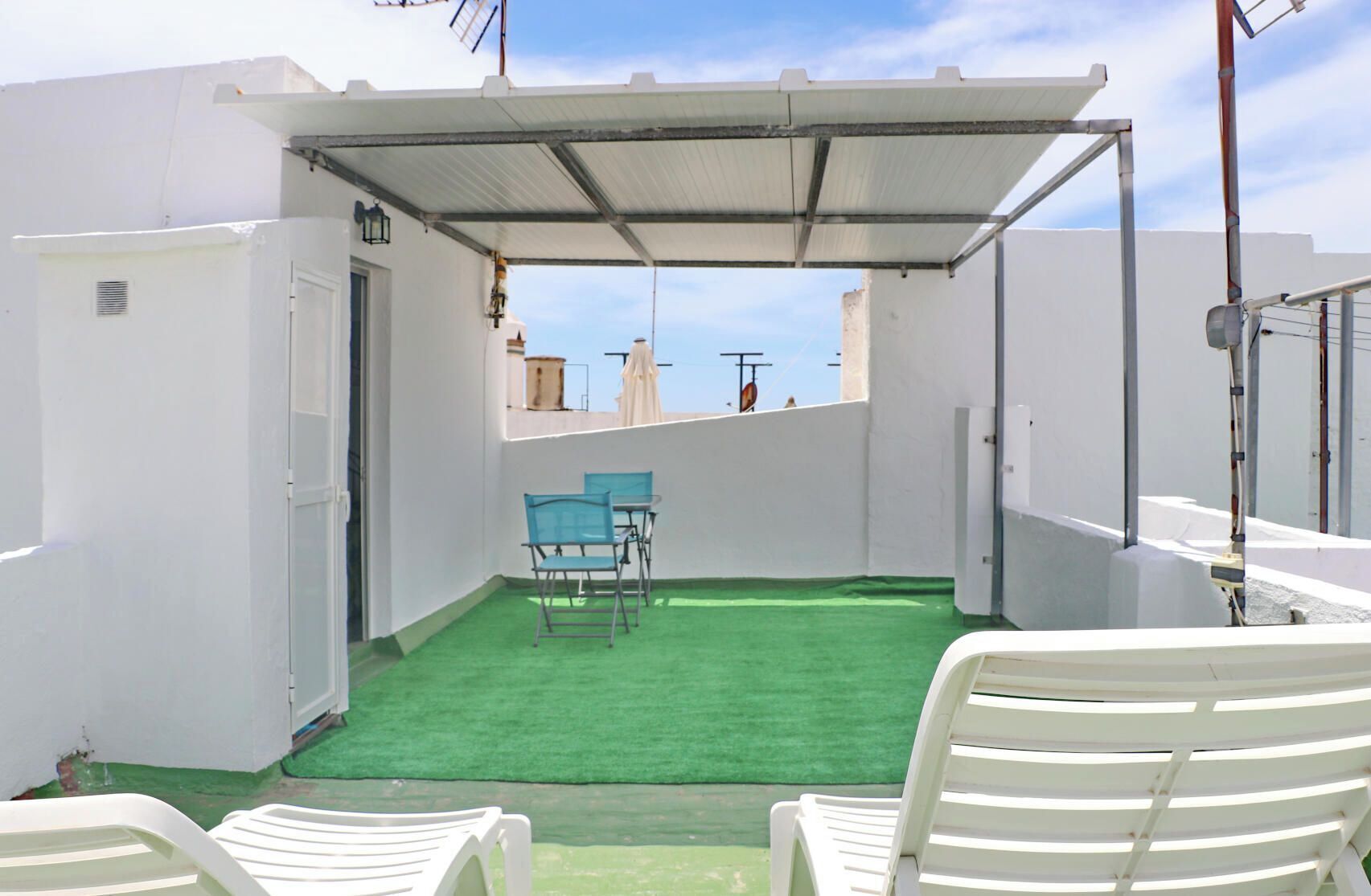 Eine Dachterrasse mit künstlichem Rasen, weißen Wänden und einer überdachten Sitzgelegenheit mit Stühlen.