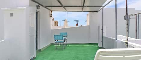 A rooftop terrace with a green artificial turf, white walls, and a covered seating area with chairs.