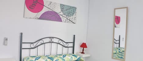 Une chambre à coucher comprenant un lit, une table de chevet, une lampe, un miroir fixé au mur et une œuvre d’art abstraite.