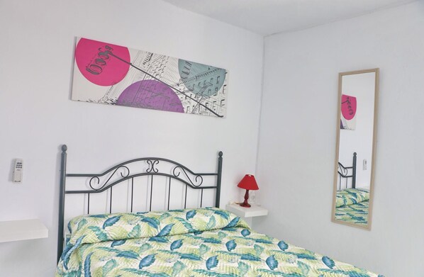 A bedroom with a bed, a nightstand, a lamp, a wall-mounted mirror, and a piece of abstract art.