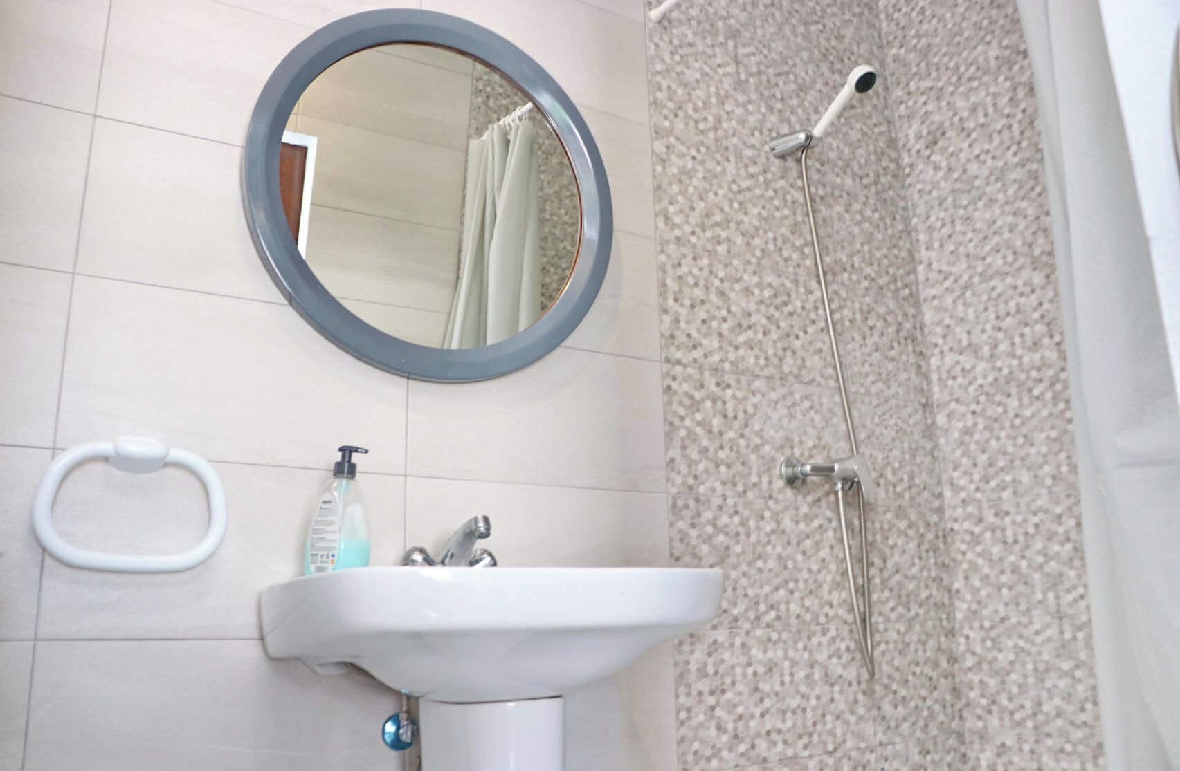 Un baño con un espejo redondo, un lavabo con dispensador de jabón, un aro para toallas y una ducha con cabezal de ducha manual.