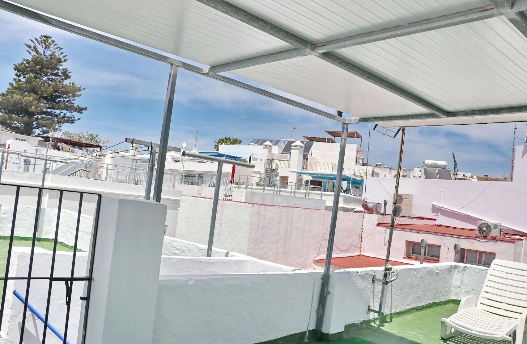 A rooftop terrace with a metal roof, white walls, and a green floor. There is a white chair and a blue railing.