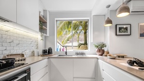 Une cuisine moderne avec des meubles blancs, une grande fenêtre donnant sur des palmiers, un four encastré et des luminaires suspendus.