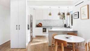 Une cuisine moderne avec des meubles blancs, un îlot central, des luminaires suspendus et un coin repas avec des chaises en bois.