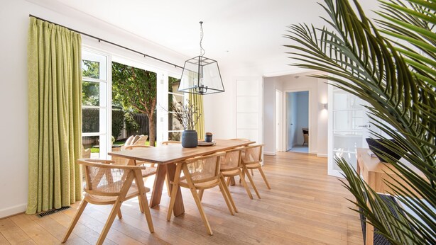 Un coin repas avec une table en bois et des chaises en osier, de grandes fenêtres avec des rideaux verts et un luminaire suspendu.